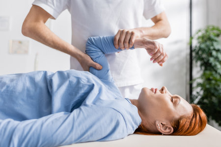 chiropractor flexing injured arm of redhead woman in rehabilitation clinicの写真素材