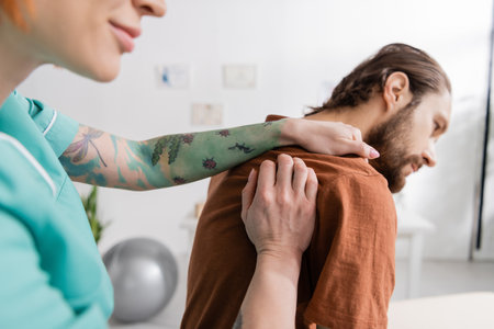 tattooed physiotherapist touching injured shoulder of bearded man in rehabilitation centerの写真素材