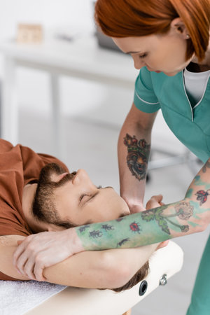 tattooed osteopath massaging injured arm of bearded man in recovery centerの写真素材