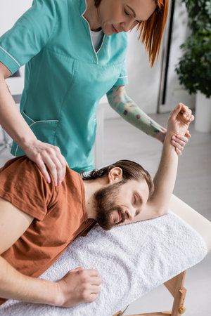 tattooed physiotherapist massaging injured shoulder of bearded man in rehabilitation centerの写真素材
