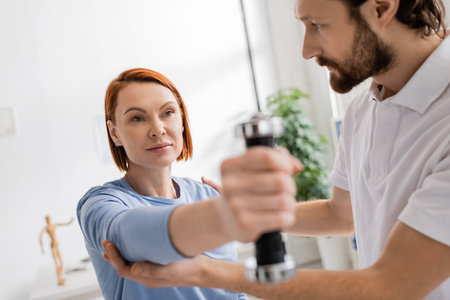 bearded physiotherapist helping redhead woman working out with dumbbell in rehab center