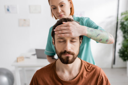 blurred physiotherapist touching forehead of bearded man with closed eyes during diagnostics in consulting roomの写真素材