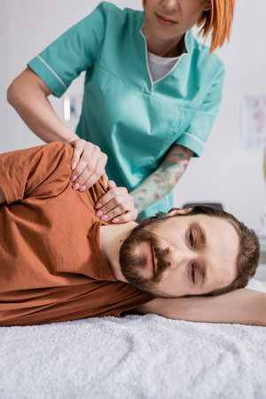 bearded man near physiotherapist massaging injured shoulder during treatment in rehabilitation centerの写真素材