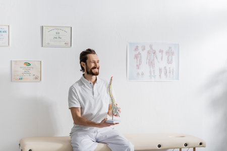 cheerful physiotherapist with spine model looking away while sitting on massage table in rehabilitation centerの写真素材