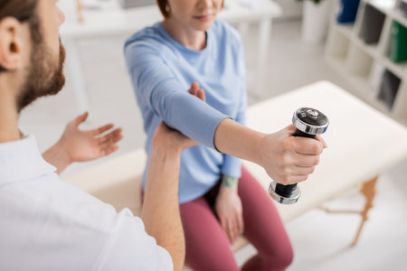 partial view of blurred woman training arm with dumbbell near rehabilitation specialist in clinicの写真素材