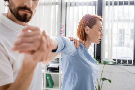 redhead woman with injured arm near blurred physiotherapist doing diagnostics in consulting roomの写真素材