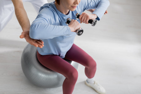 partial view of woman sitting on fitness ball and working out with dumbbells near rehabilitologist in clinicの写真素材