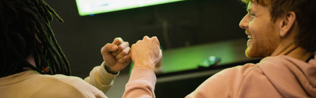 Smiling young man doing fist bump with multiracial friend in gaming club, bannerの写真素材