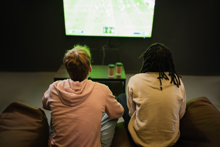 Back view of interracial friends watching sports match near blurred coffee to go in cyber club