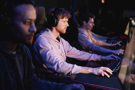Young man in headphones playing video game on computer near blurred interracial team in gaming club