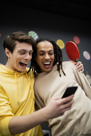 Positive interracial friends with tennis racket using mobile phone in gaming clubの写真素材