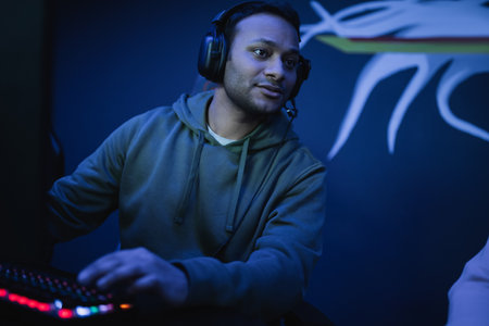 Indian gamer in headphones looking away near keyboard in cyber club