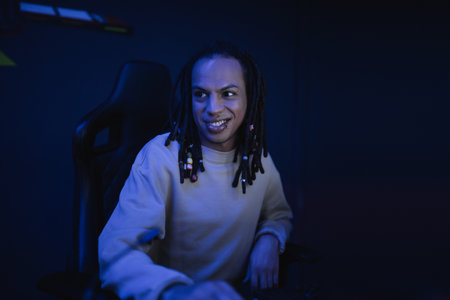 Smiling multiracial gamer with dreadlocks sitting in cyber club with lighting