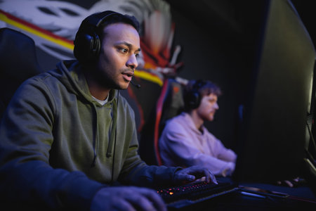Indian player in headphones playing computer game with blurred friend in gaming clubの写真素材