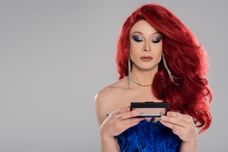 Elegant drag queen in red wig holding eye shadow isolated on greyの写真素材