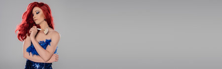 Well dressed drag queen in red wig and dress holding cosmetic brush isolated on grey, bannerの写真素材