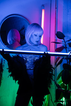 Stylish drag queen in top and gloves holding neon lamp at homeの写真素材
