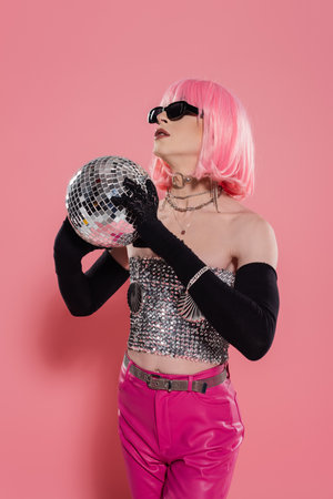 Stylish transgender person in sunglasses and gloves holding disco ball on pink backgroundの写真素材