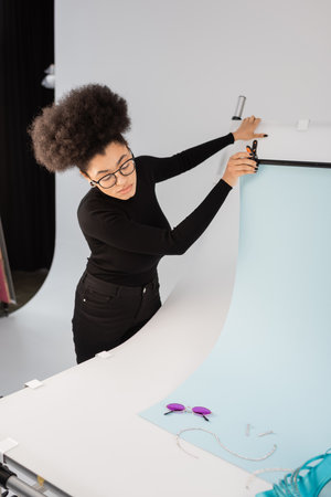 african american content maker adjusting background sheet near trendy sunglasses and jewelry on...