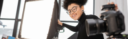cheerful african american content producer in eyeglasses looking at camera near spotlight and blurred digital camera, bannerの写真素材