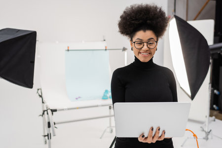 happy african american content producer with laptop smiling at camera near reflectors and shooting...