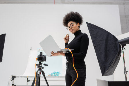 african american content maker with laptop smiling near softbox and digital camera in contemporary...