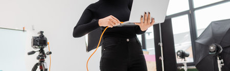 partial view of african american content maker plugging cable in laptop while working in photo...