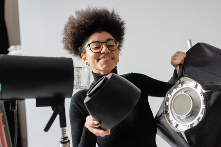 carefree african american content producer holding softbox near strobe lamp and smiling at camera in...
