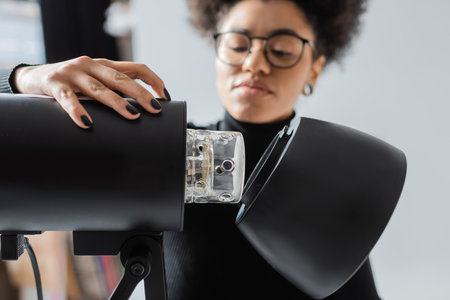 blurred african american content producer in eyeglasses assembling strobe lamp in photo studioの写真素材