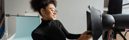 pleased african american content maker in black turtleneck and eyeglasses working with strobe floodlight in photo studio, bannerの写真素材