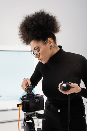 stylish african american content manager holding exposure meter and adjusting digital camera in...