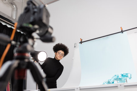 african american content maker looking at blurred digital camera near spotlight and shooting table...