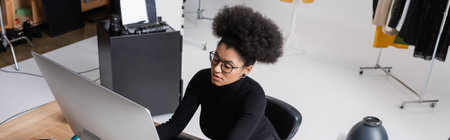 high angle view of african american content manager in eyeglasses and black turtleneck working on computer in photo studio, bannerの写真素材