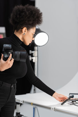 african american content producer with digital camera holding cosmetic brush near cosmetics on shooting table in photo studioの写真素材