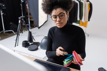 african american retoucher in black turtleneck and eyeglasses holding color samples and looking at camera near graphic tablet in photo studioの写真素材
