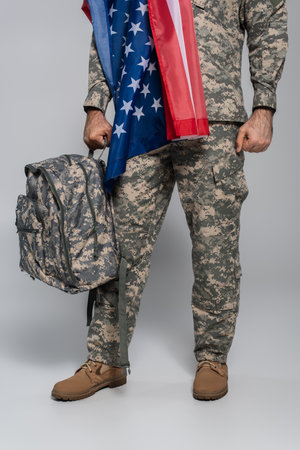 cropped view of army soldier in uniform with flag of United States of America holding backpack on greyの写真素材