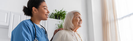 cheerful multiracial nurse in blue uniform looking at happy senior woman with grey hair at home,...