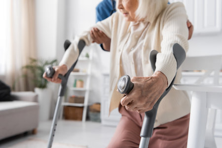 cropped view of multiracial caregiver in uniform helping senior woman using crutches to walk at homeの写真素材