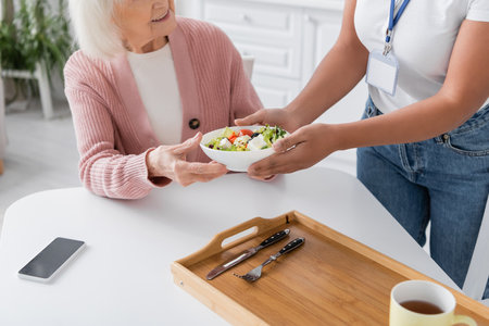 cropped view of multiracial social worker giving bowl with salad to happy senior womanの写真素材