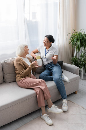 happy multiracial social worker chatting with senior woman while having tea in living roomの写真素材