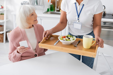 multiracial social worker holding tray with lunch for happy senior woman with grey hairの写真素材