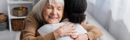 happy senior woman with closed eyes hugging multiracial social worker in living room, bannerの写真素材
