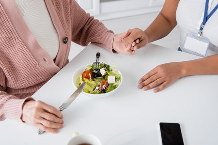 top view of senior woman holding hands with multiracial caregiver next to lunch on tableの写真素材