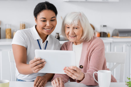 joyful multiracial social worker holding digital tablet near senior woman in kitchenの写真素材