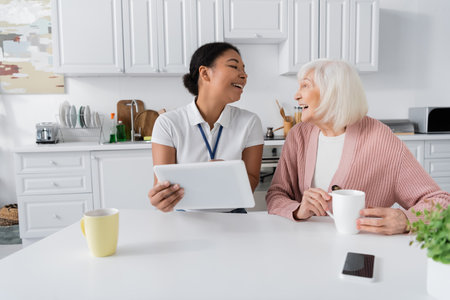 cheerful multiracial social worker holding digital tablet near senior woman in kitchenの写真素材