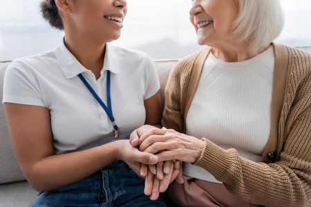 cropped view of happy multiracial social worker holding hands with senior woman in living room