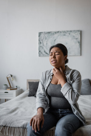 Stressed multiracial woman touching sore throat while sitting on bed at homeの写真素材