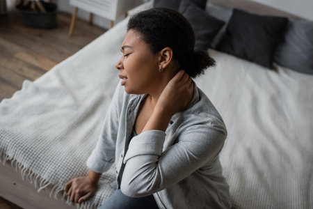 Side view of upset multiracial woman with depression touching neck while sitting on bedの写真素材
