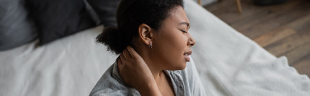 Side view of sad multiracial woman touching neck near bed at home, bannerの写真素材
