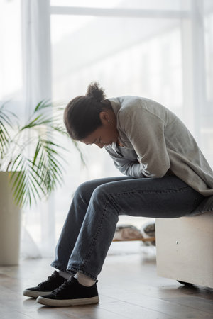 Sad multiracial woman feeling unwell while sitting on bed at home